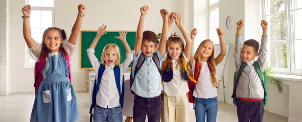 Small team of children having fun at school. Group portrait of happy classmates with backpacks standing in row in the classroom, smiling, raising arms and shouting Yay, Hooray. Back to school concept
