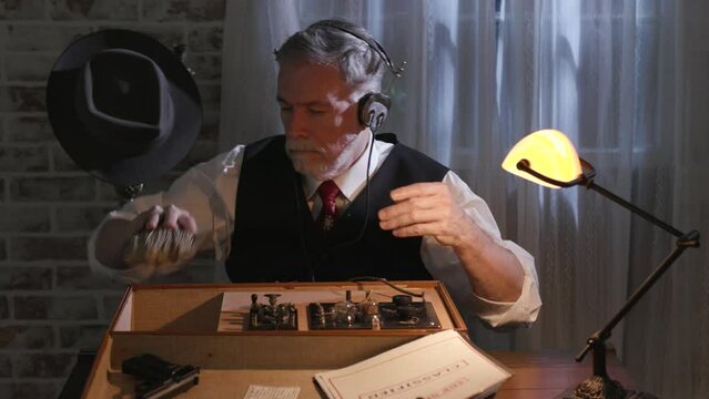 A man on assignment during WWII finishes transmitting messages from a wireless radio transmitter hidden inside a small suitcase and begins putting things back in place.