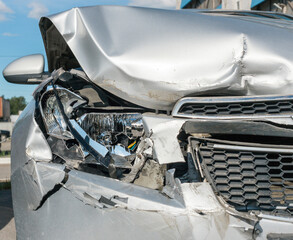 The front part of the silver car is damaged as a result of an accident on the road. The front part of the car is silver, damaged and broken as a result of an accident. Hood, headlight, bumper. 
