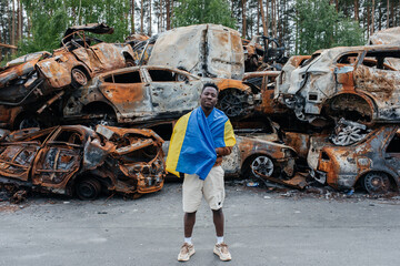 African man with Ukrainian flag stands against cars cemetery in Irpin. Consequences of the Russian...