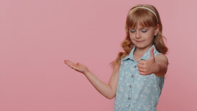 Toddler toddler girl showing thumbs up pointing empty place, advertising area for commercial text, copy space for goods promotion. Young little child kid. Studio shot indoors on pink wall background