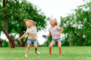 Two little girls sisters dance in the summer in the park on the grass and hold a mobile phone....