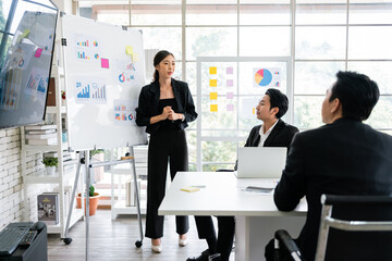 A cheerful and confident Asian businesswoman stands, present bar charts data from a whiteboard to her office colleagues. Asian business women leader role at the meeting.