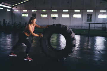 Obraz na płótnie Canvas Slim female bodybuilder using wheel equipment during crossfit practice in sportive gym studio, Caucaisan woman trainer in tracksuit lifting tire during time for hard exercising and slimming