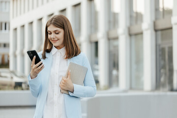 Smiling caucasian woman wearing formal suit using mobile phone typing text messages walking holding...