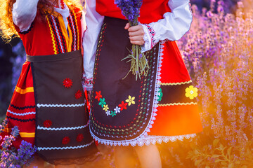 Beautiful girls in traditional Bulgarian folklore costumes in lavender field during harvest and sunset - obrazy, fototapety, plakaty