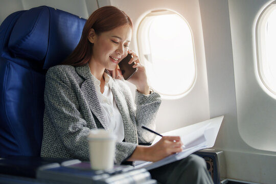 Portrait Of A Successful Asian Business Woman Or Female Entrepreneur In Formal Suit In A Plane Sits In A Business Class Seat And Uses Smartphone With Documents For Work During Flight