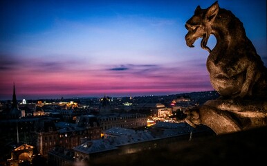 Closeup shot of a dragon sculpture on Notre Dame over a background of the city and the colorful sky