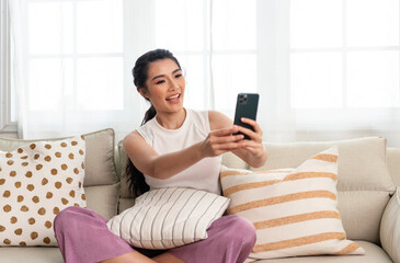 Relaxed smiling asian woman on the couch at home, she is using a smartphone for video call.