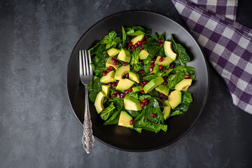 Avocado salad with spinach and pomegranate in a black plate on black background.