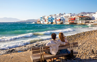 Romantic young couple sitting against the waves, Mykonos island, Greece, famous Little Venice area...