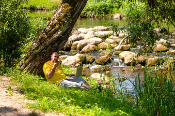 man with laptop and smartphone working outside in the meadow by the river, outdoor office concept