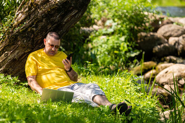 Naklejka na ściany i meble man with laptop and smartphone working outside in the meadow by the river, outdoor office concept