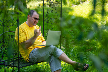 mature man with laptop working outside in garden and show fucking gesture into monitor