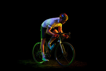 Fototapeta premium Portrait of young male cyclist on bicycle in cycling shorts and protective helmet isolated on dark background in neon. Concept of sport, speed, energy