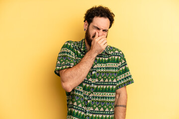 young adult bearded man feeling disgusted, holding nose to avoid smelling a foul and unpleasant...
