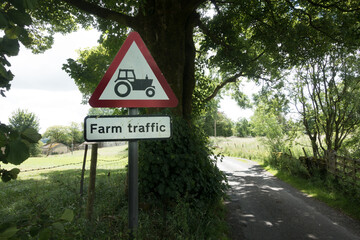 A red triangular road sign has a tractor symbol and words farm traffic situated on a country lane