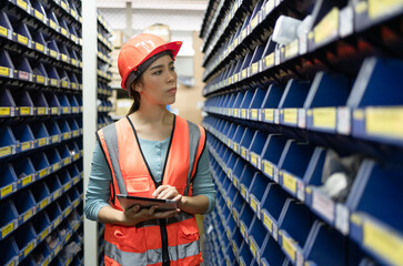 Young Asian female employee worker in safety vest and helmet using tablet checking products spare...