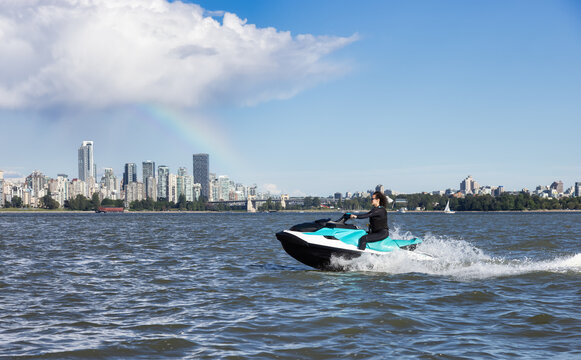 Adventurous Caucasian Woman on Sea-Doo riding in the Ocean. Modern City in background. Downtown Vancouver, British Columbia, Canada. Colorful Rainbow