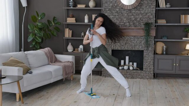 Funny young hispanic woman housewife cleaning at home washes floor in living room cheerful excited girl expressively sings uses mop as microphone performs movement energetic shaking long curly hair
