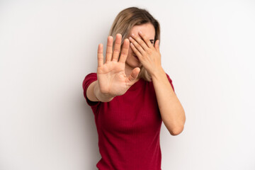 young adult blonde woman covering face with hand and putting other hand up front to stop camera,...