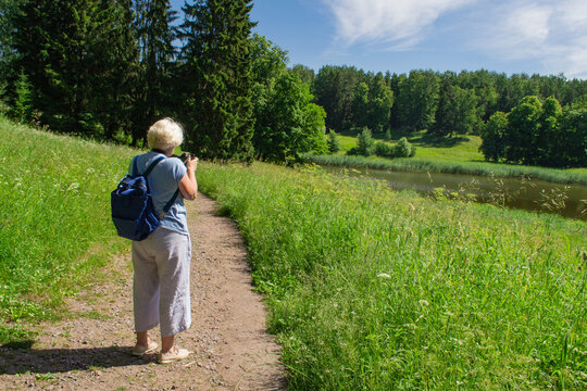 An elderly woman with gray hair in a blue T-shirt and light trousers with a backpack on her back photographs a beautiful summer landscape with a professional camera. Soft focus, leisure, hobby, nature