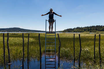 Young woman with hat standing on ladder on educational trail Olsina from back. Sumava reserve,...
