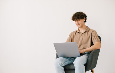 Happy young man using laptop while sitting in armchair against white wall, copy space. Millennial male communicating online, working or studying remotely on portable pc