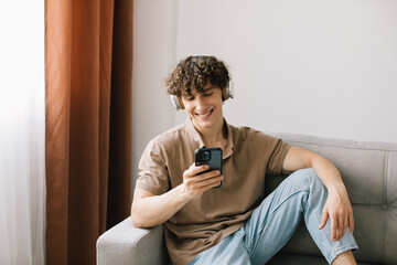 Portrait of young happy curly haired man using smartphone with headphones while sitting on sofa in living room and resting