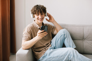 Portrait of young happy curly haired man using smartphone with headphones while sitting on sofa in living room and resting