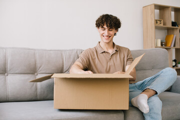 Happy young smiling curly man opening box with ordered goods gifts, presents at home on couch....