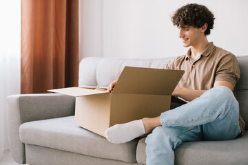 Happy young smiling curly man opening box with ordered goods gifts, presents at home on couch. Online shopper male customer opening online shop parcel.