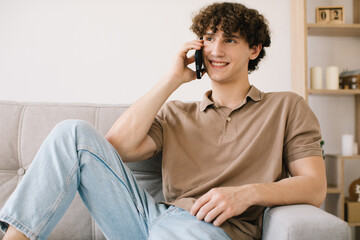 Obraz premium Attractive curly hair young male student talking by phone while sitting on a sofa in living room