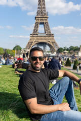 young man sitting on the lawn wearing sunglasses with the eiffel tower in the background, in Paris.