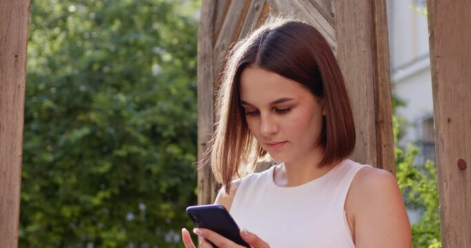 Portrait of beautiful happy young woman with short dark hair texting mesage on smartphone and smiling to camera. Pretty cheerful female tapping and typing on mobile phone outside on sunny day.