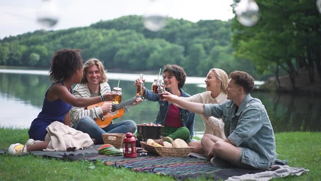 Group of friends having fun on picnic near a lake, sitting on blanket eating and playing guitar.