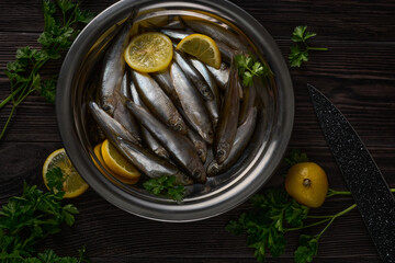 In a metal plate, which stands on a dark wooden table, there is a lot of small fish. Greens and lemons lie next to the plate.