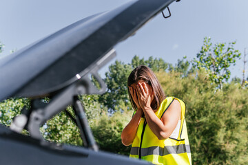Young girl in yellow reflective vest with her hands on her face when she sees the serious breakdown of her vehicle. Woman in front scared to see that her car is broken and has to call for assistance.