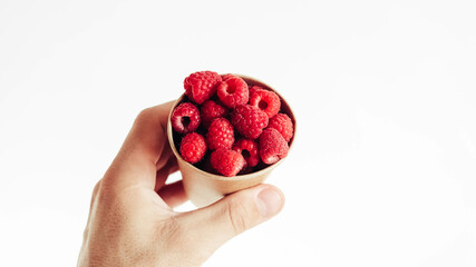 Fresh raspberries in a paper cup held by a male hand on a white background