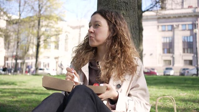 Woman eats delicious cake with fresh fruits using plastic fork near tree against buildings. Young lady enjoys lunch in city public garden slow motion