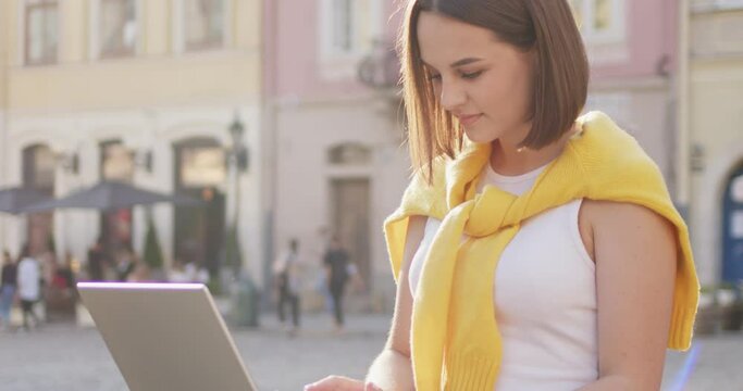 Young Caucasian beautiful female freelancer working on laptop computer on sunny day while sitting outdoors in city square. Stylish woman texting and typing on keyboard while mailing online.