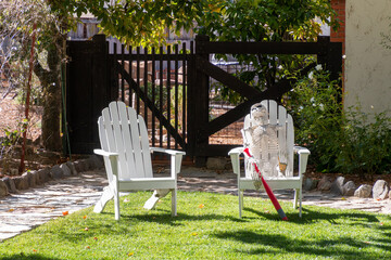 Halloween. A skeleton is siting in a white outdoor chair. It is holding a red plastic baseball bat. Another white chair is next to the other chair. They are on a green lawn. A gate is behind them.