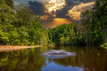 a still lake with a water fountain in the center surrounded by lush green trees and grass reflecting off the water with powerful clouds at sunset at Dupree Park in Woodstock Georgia USA - obrazy, fototapety, plakaty