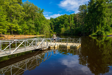 a man wearing a white shirt and a backpack fishing on a dock with a metal hand rail on a lake surrounded by lush green trees at Dupree Park in Woodstock Georgia USA - Powered by Adobe