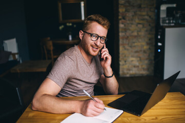 Smart caucasian business man calling via smartphone while sitting indoors front laptop computer. Redhead male freelancer working online on netbook while having mobile conversation on cellphone