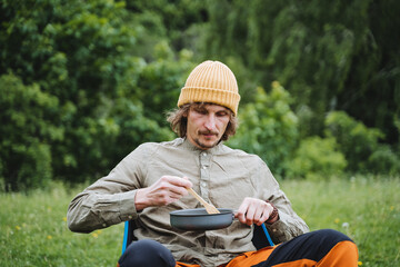 Fototapeta premium A person on a hike eats food from a frying pan, tourist food in nature, a guy on a hike has breakfast with scrambled eggs, a summer vacation in nature camping