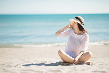 Beautiful teenager teen girl sitting on the beach, wearing white beachwear and straw hat. Summer holidays and vacation concept.