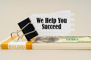 On a gray surface is a pack of dollars on which there is a clip with torn paper and the inscription -We Help You Succeed