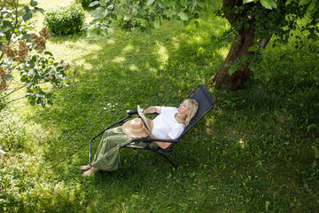 Woman reading a book in garden recliner chair. Summer vacation in the yard. Mature Scandinavian...