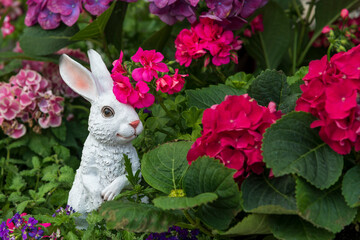 A figurine of a white rabbit stands in a flower bed near bright colorful geranium flowers. Copy space for your text. Easter decoration theme. - obrazy, fototapety, plakaty